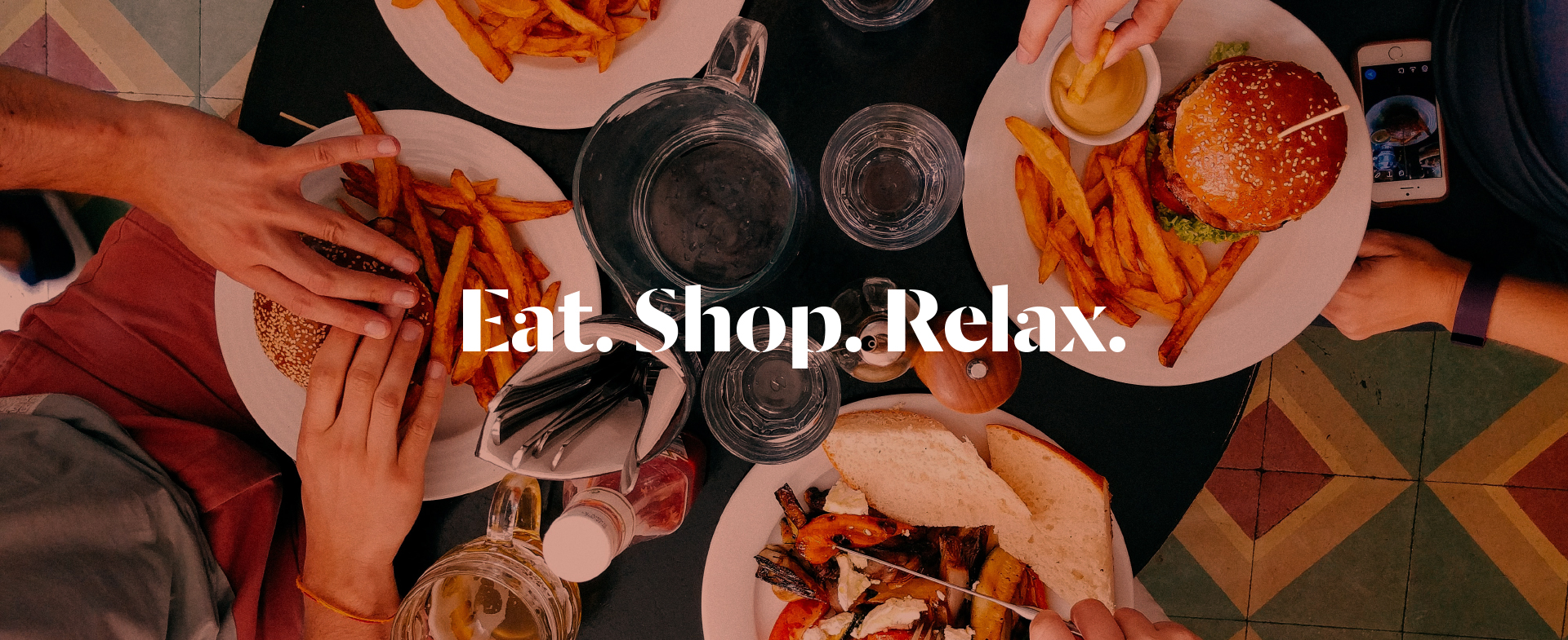A photo taken from above of people eating and drinking. The table is laid with plates of burgers and chips and glasses of water. Text over the image says Eat. Shop. Relax.