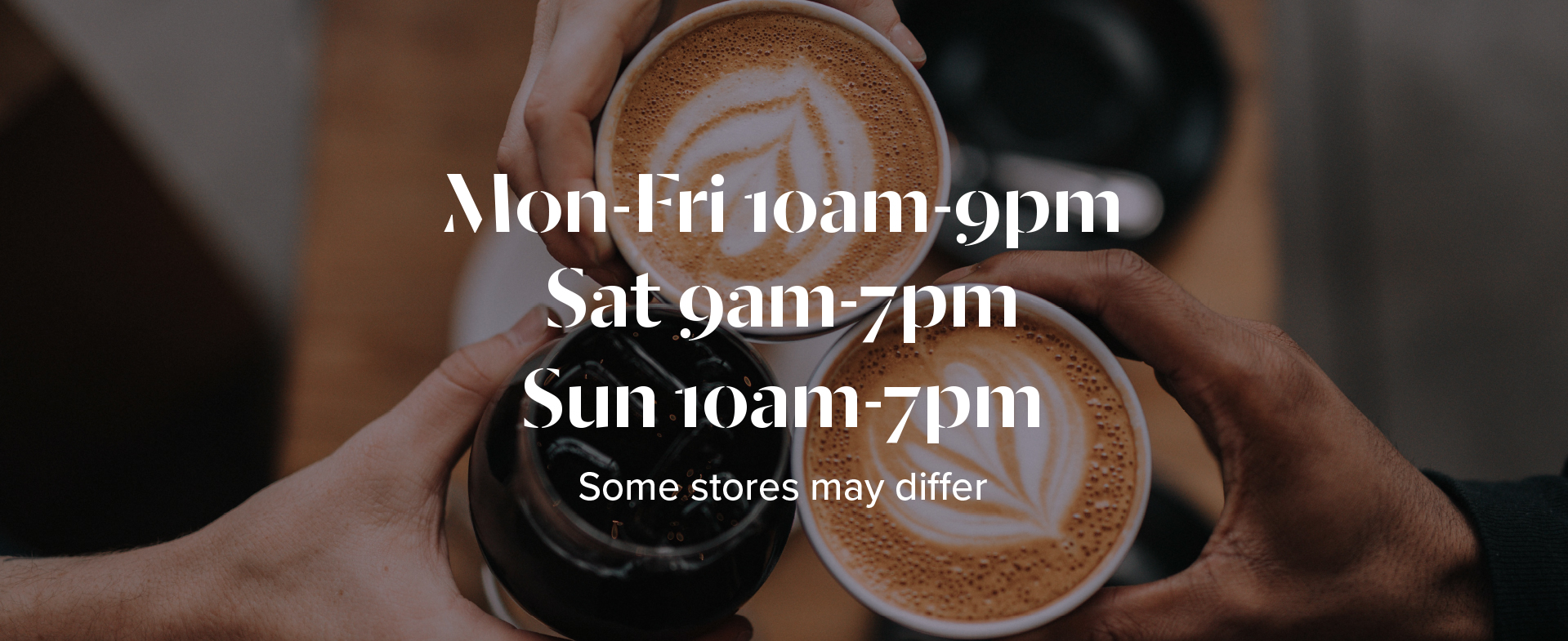 A close up photo taken from above of 3 people holding drinks in a motion of cheers. Two cups contain coffees with latte art and the third is a cold drink with ice. Text over the image contains store opening times.