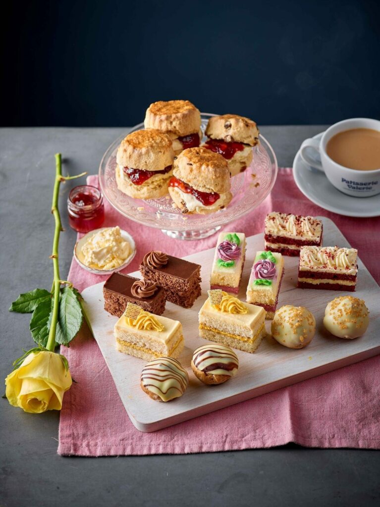 A Patisserie Valerie afternoon tea cake spread including scones next to a yellow rose and a cup of tea.